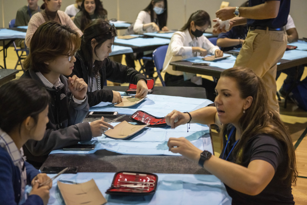 An instructor is teaching three students how to suture with scissors, needle, and suturing pad.
