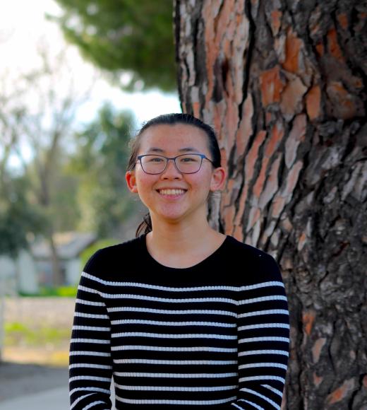 Smiling woman in glasses, black sweater, standing by a tree.