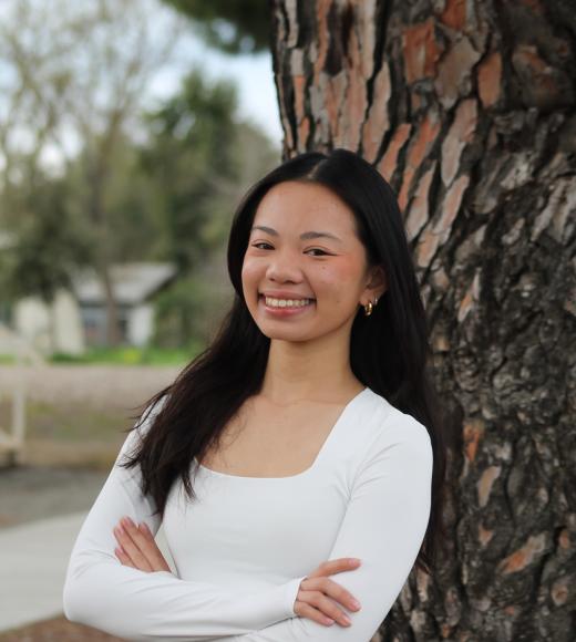 Young woman in a white sweater vest stands by a tree, looking joyful.