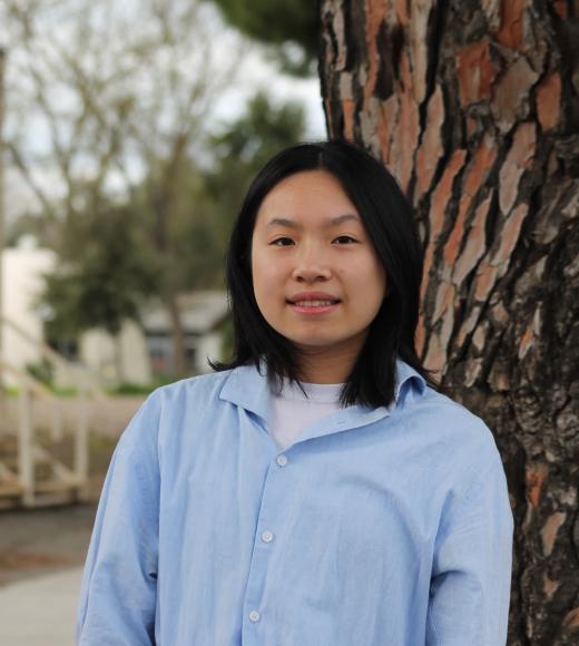 Young woman in a blue shirt stands by a tree, looking joyful.