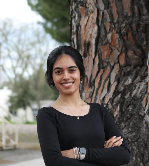 Young woman in a black sweater stands by a tree, looking joyful.