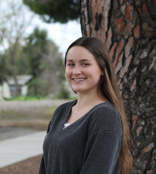 Young woman in a black sweater stands by a tree, looking joyful.