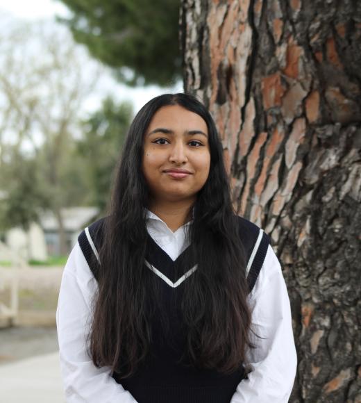 Young woman in a black sweater vest stands by a tree, looking joyful.