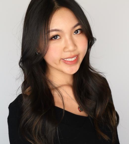 Young woman with long, wavy black hair smiles, wearing a black top against a light background.