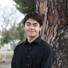 Young man in a black shirt stands by a tree, looking joyful.