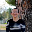 Smiling woman in glasses, black sweater, standing by a tree.