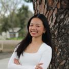 Young woman in a white sweater vest stands by a tree, looking joyful.
