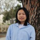 Young woman in a blue shirt stands by a tree, looking joyful.