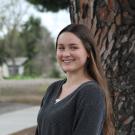 Young woman in a black sweater stands by a tree, looking joyful.
