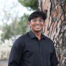 Young man in a black shirt stands by a tree, looking joyful.