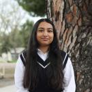 Young woman in a black sweater vest stands by a tree, looking joyful.