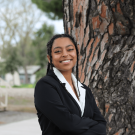 Young woman in a black sweater stands by a tree, looking joyful.